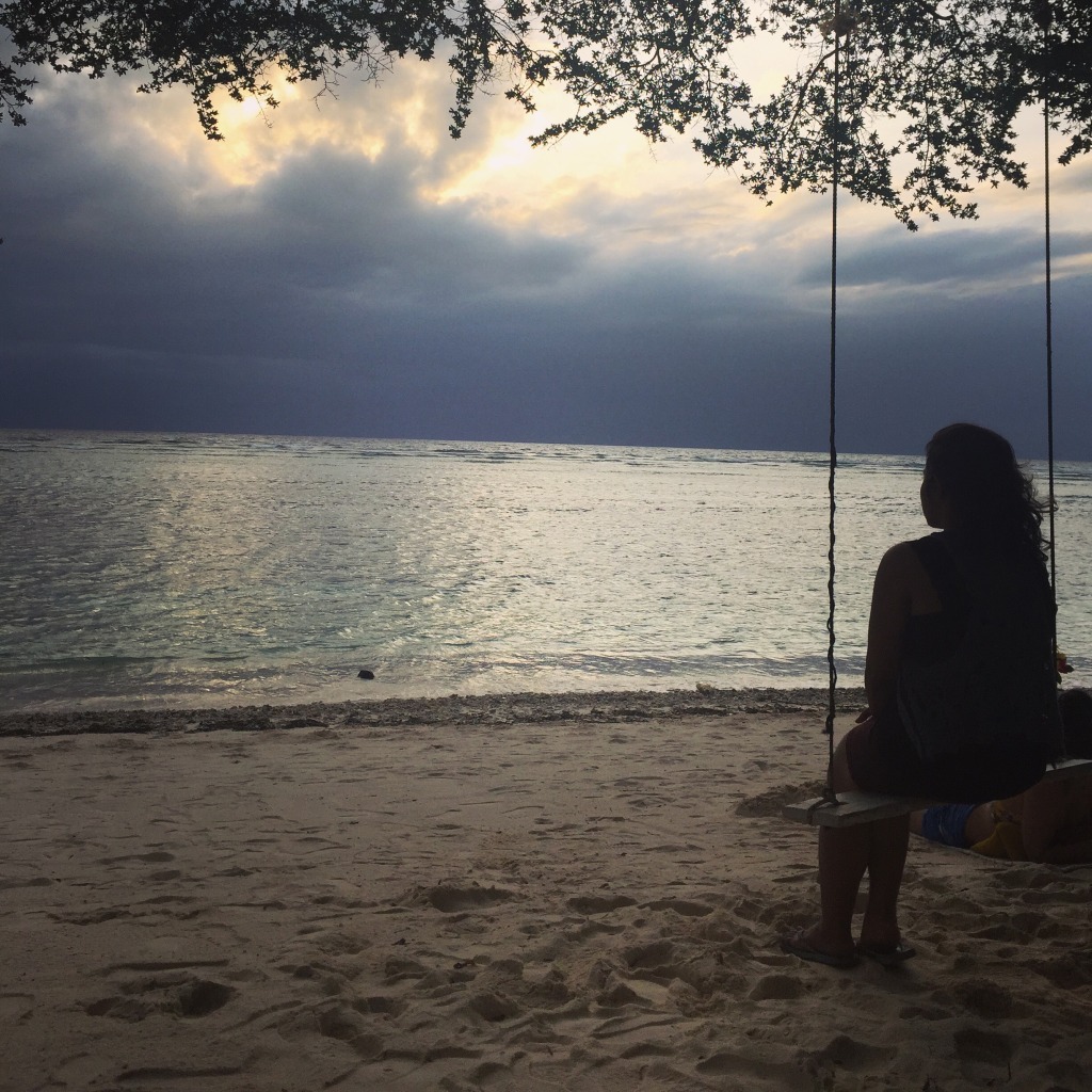 The author, on a swing, gazing at a large body of water, her back to the viewer, in Indonesia.