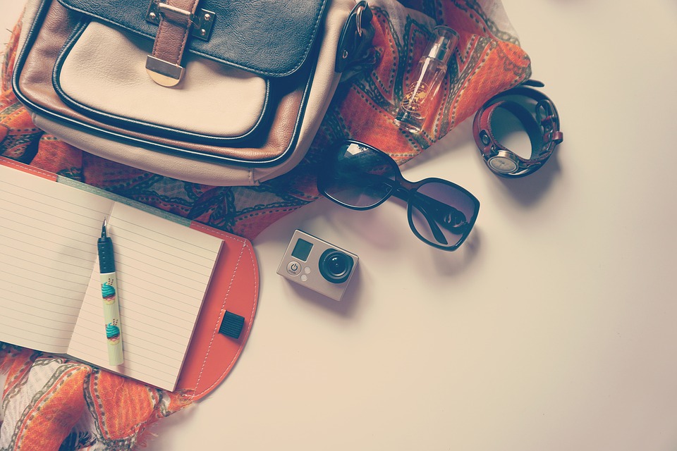 Travel bag with contents spilled out: sunglasses, notebook and pen, camera, a watch, and perfume.