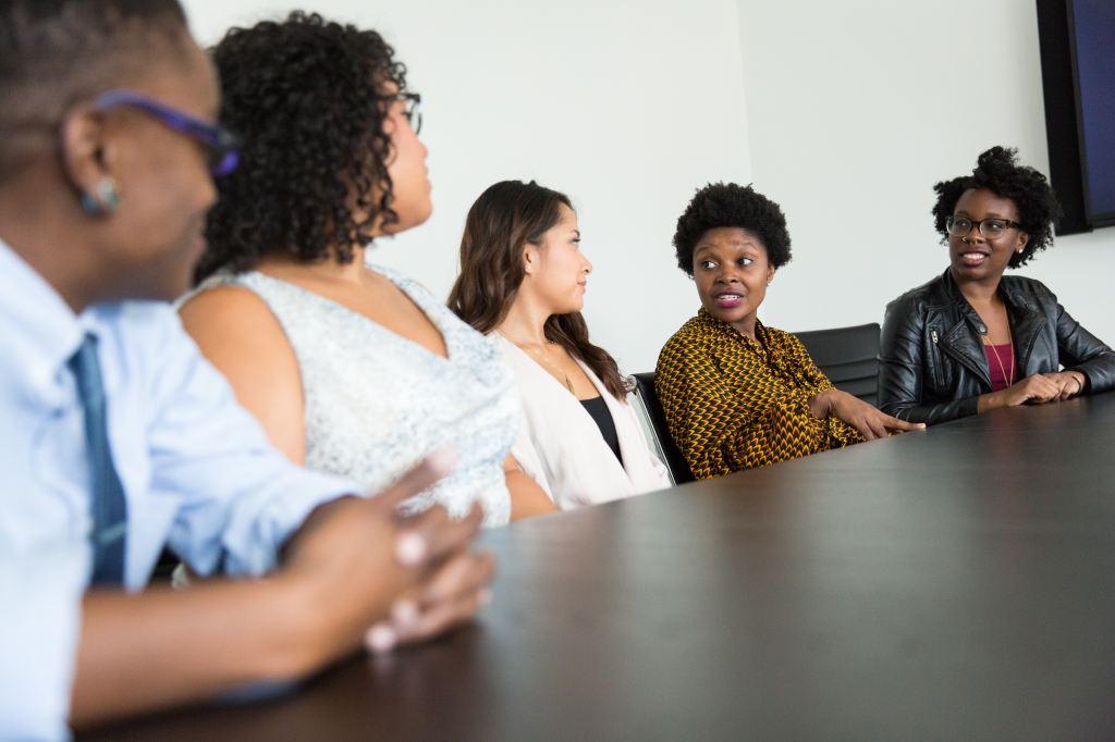 five women of colour sitting around boardroom table, talking