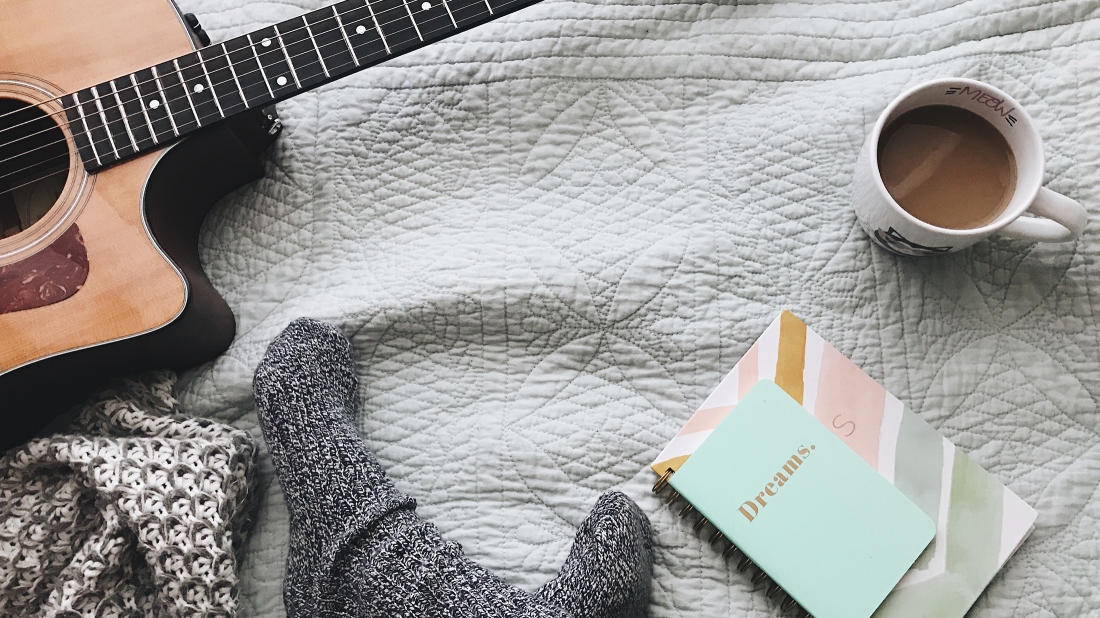 Mug of coffee on a bedspread, with a guitar, a couple of notebooks (the top one has "Dreams" written on the cover), and a couple of feet in socks poking from bottom of image.