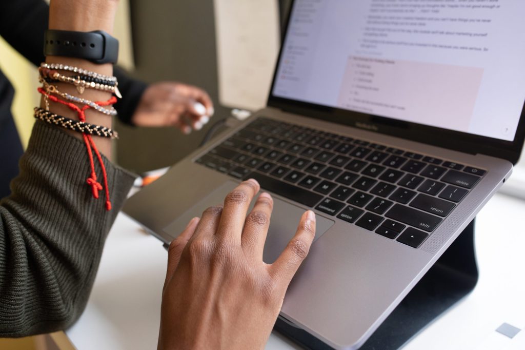 Hand in foreground with finger on trackpad of MacBook Pro. Screen of laptop partly visible. Other hand raised. Many colourful braided bracelets and black watch band seen on raised hand.