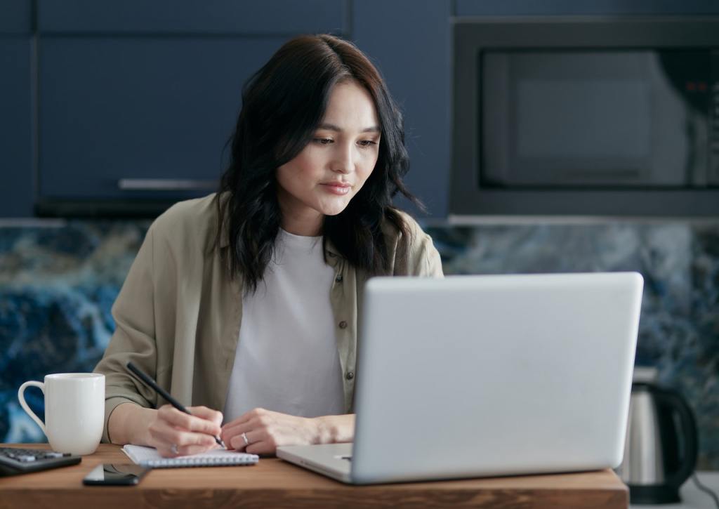 A woman looking at her laptop and writing with a pen in a notebook.