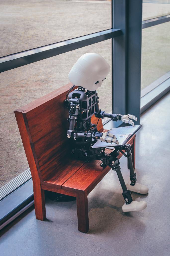 A mechanical person (robot) sits on a bench and reads a book. 