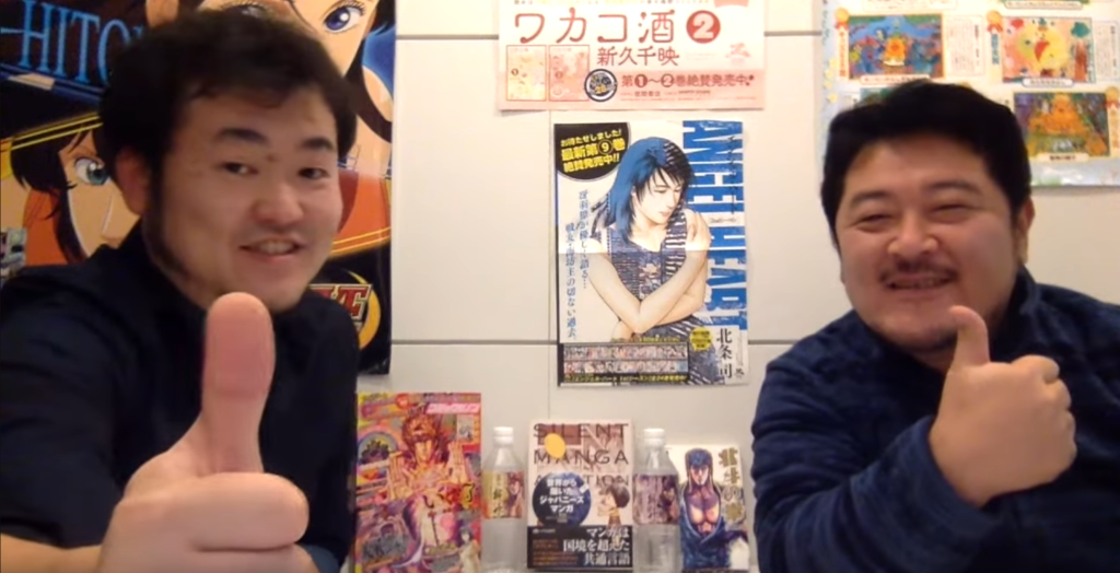 A photo of Shuichi Mochida and Taiyo Nakashima giving the thumbs up, with manga art on the wall behind them. The photo is taken from one of their YouTube videos 