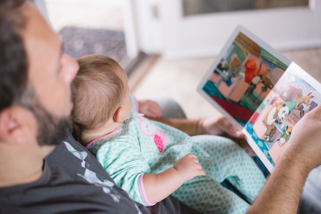 A man reads a picture book to a baby that is reclining on his lap.