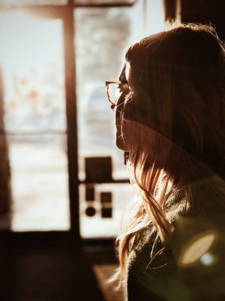 A woman sitting in a darkened room looks out into the light through a large window.