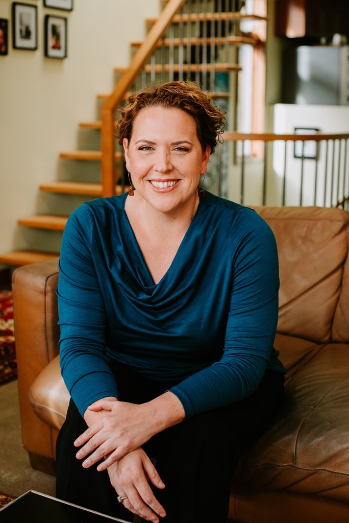 Lorie Boucher is seated on a sofa. A staircase is in the background with framed pictures hanging on the wall. 