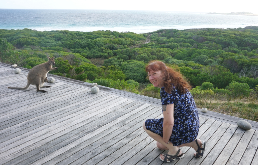 Gael Spivak is pictured outdoors, 10 feet away from a wallaby. Lush foliage and a large body of water can be seen in the distance.