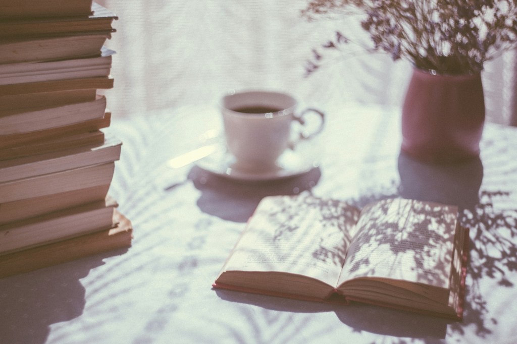 A photo of an open book with a cup of coffee, a vase of flowers, and a stack of books laying on a table.