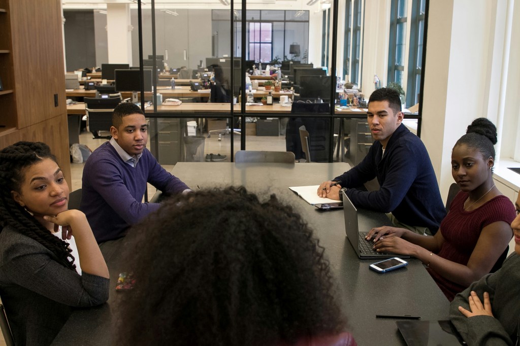 A group of people sit around a boardroom table listening to a speaker at the head of the table.