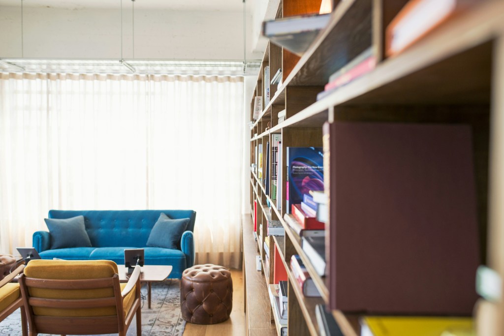 A full bookshelf beside a blue sofa and a yellow reading chair. All three are in front of a brightly lit window.