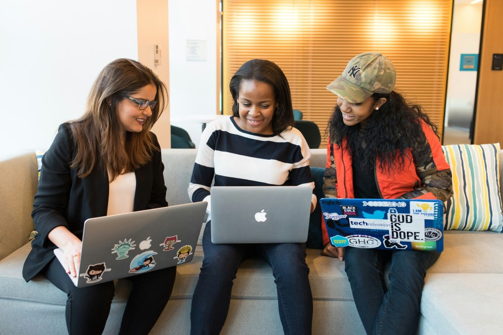 Three individuals sit on a sofa, with three open laptops look at the laptop screen in the middle of the group.