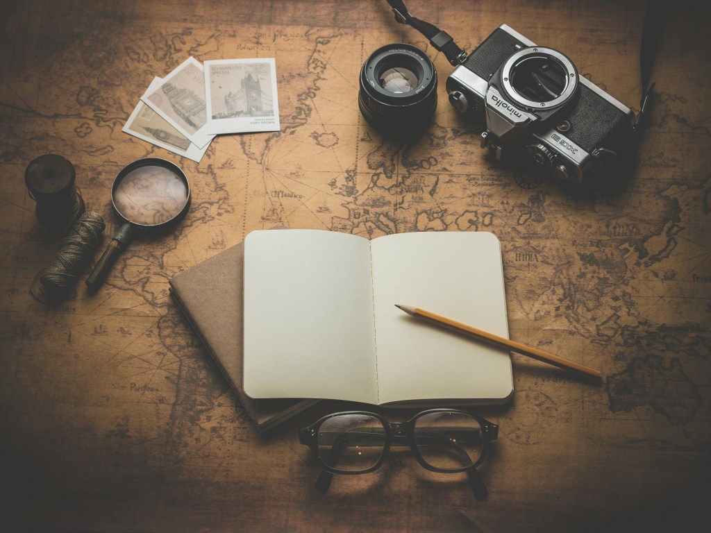 An open, blank notebook sits on top of a world map. A pair of black-framed glasses are located below the notebook. A yellow pencil sits on the notebook. An old-fashioned camera, a magnifying glass, and polaroid photos are above the notebook.