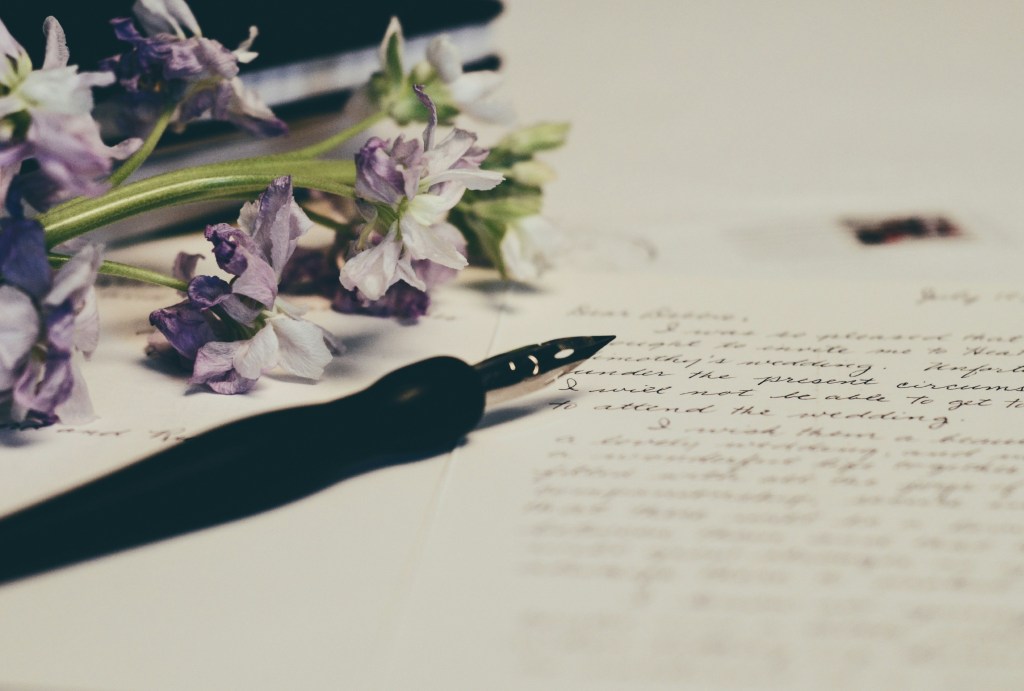 An old fashioned pen sit beside a handwritten letter. Purple flowers are on the upper left-hand corner by the pen and paper.