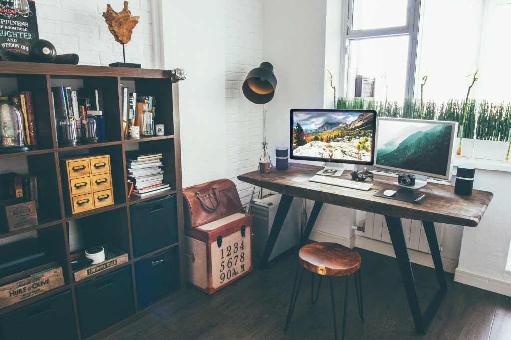 A rustic office with a desk, a stool, several computer monitors, and a wide bookcase.
