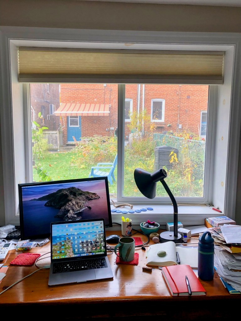 A desk with computer monitor, laptop, reading lamp and notebooks.