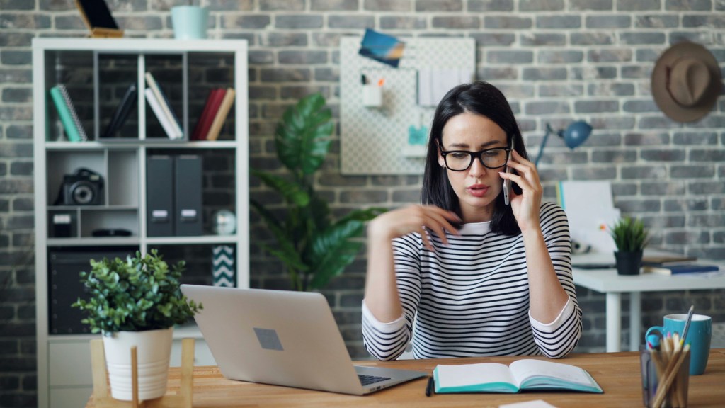 A woman sitting in front of a laptop and speaking on a cellphone.