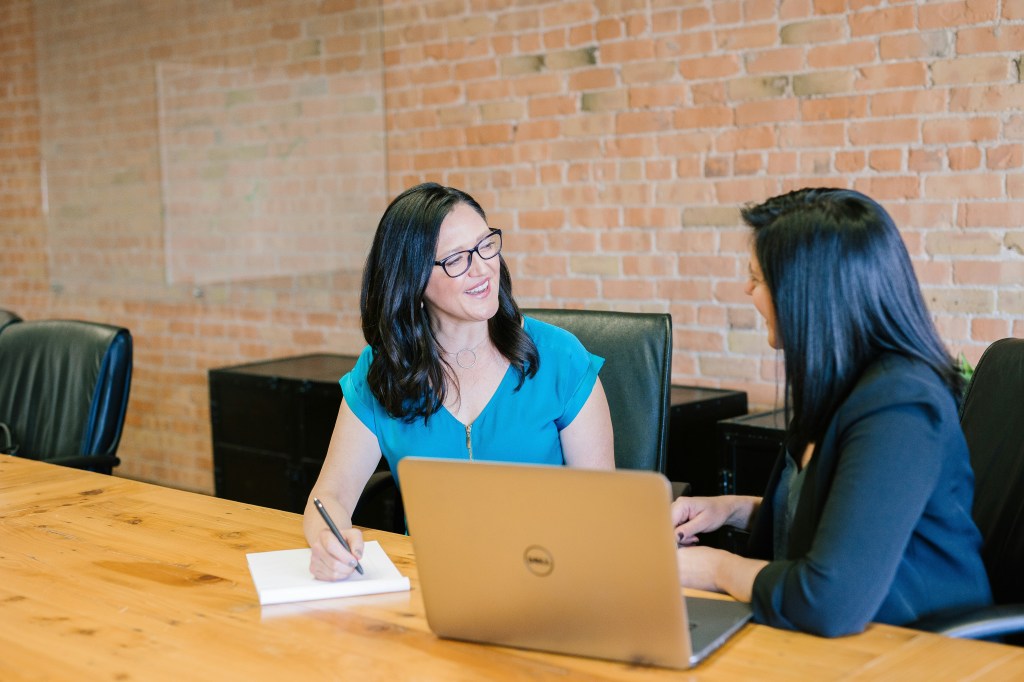 Two women sitting at a table; one with a laptop and the other with a notebook and pen.