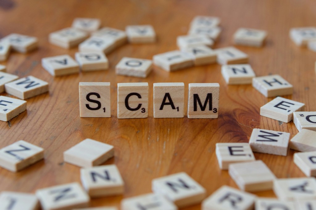 Scrabble tiles scattered on a table with the word "SCAM" spelled out in upright tiles in the centre.