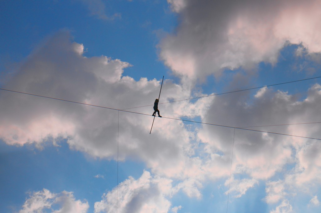 Figure photographed from below balancing on a tightrope, with clouds in the background.